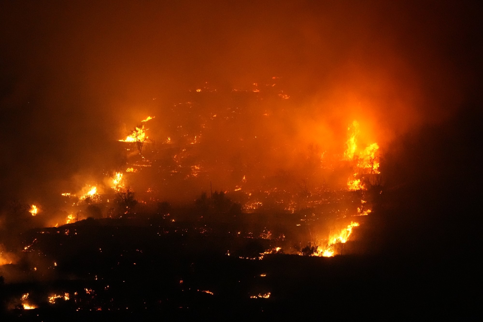 Photo of a wildfire taken at night. Orange flames, lots of embers and smoke.