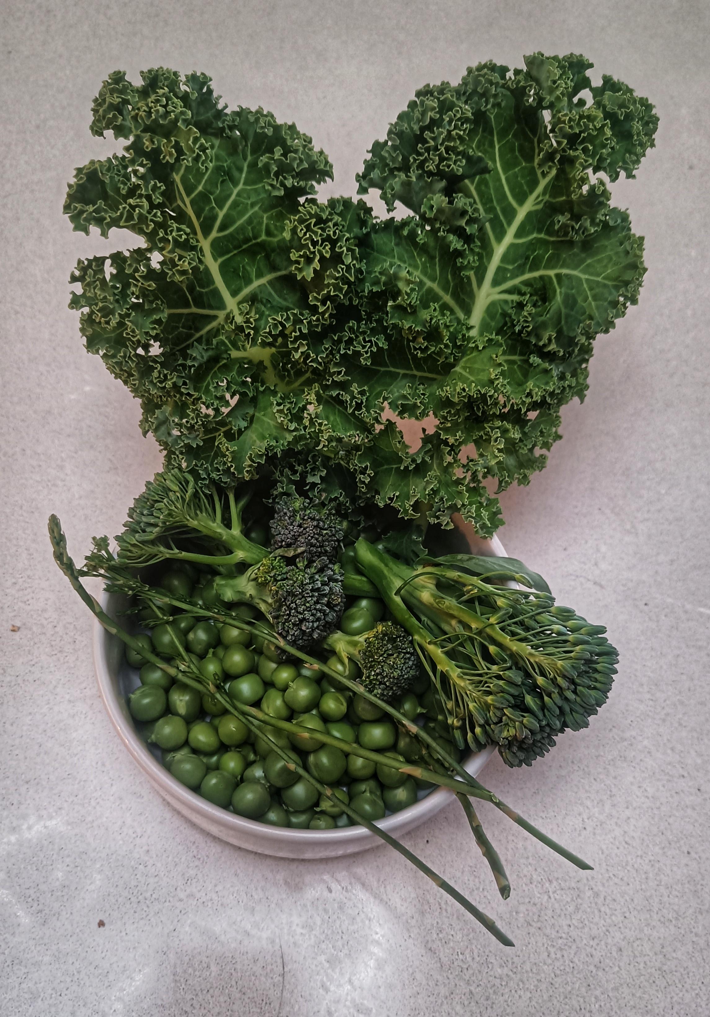 A picture showing freshly picked peas, asparagus, two types of broccoli and curly kale. All are green, in a white bowl on a white worktop.