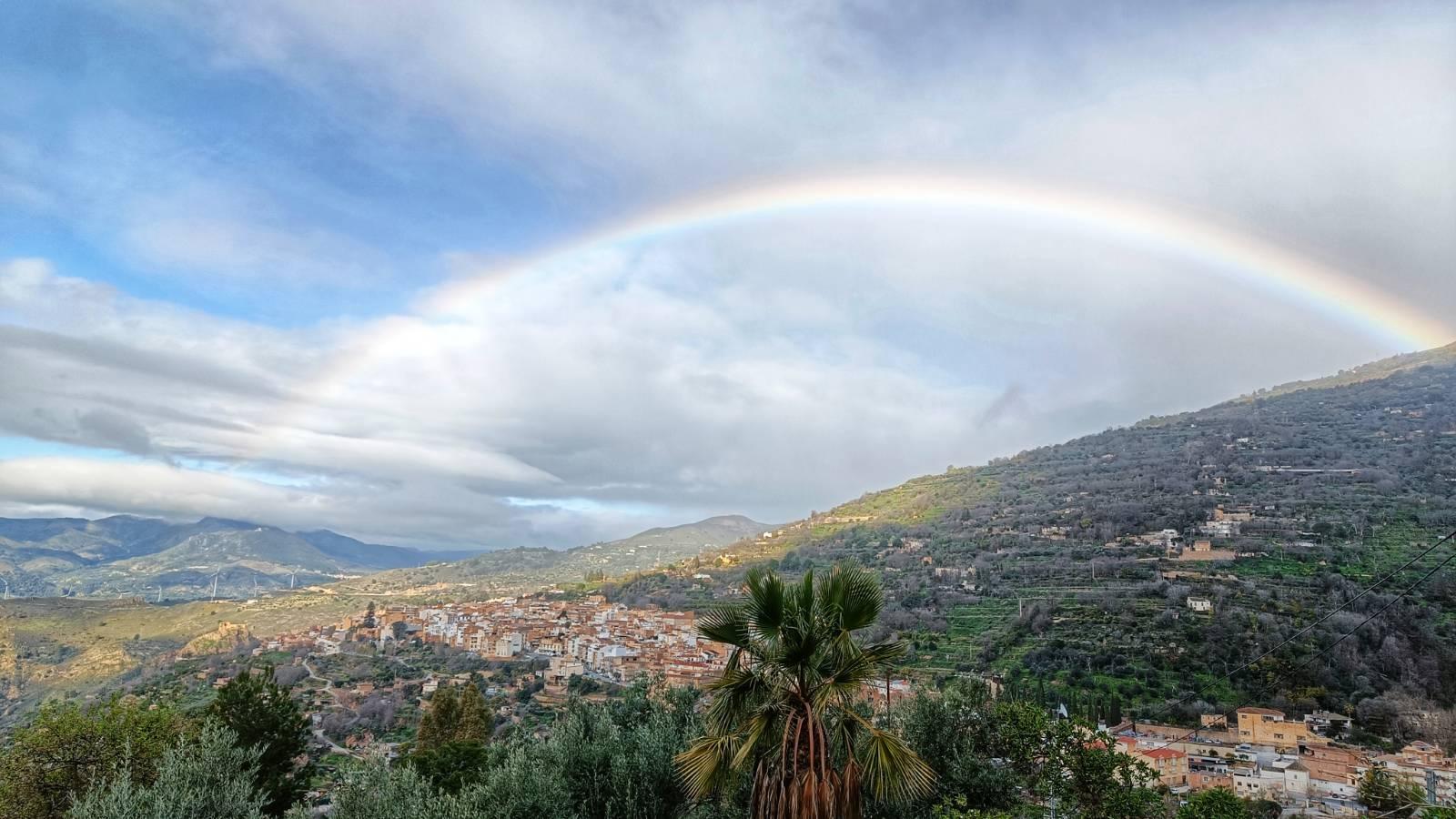 A picture showing a rainbow arching through a lightly clouded sky over a Spanish village set on a hillside with many green trees.
