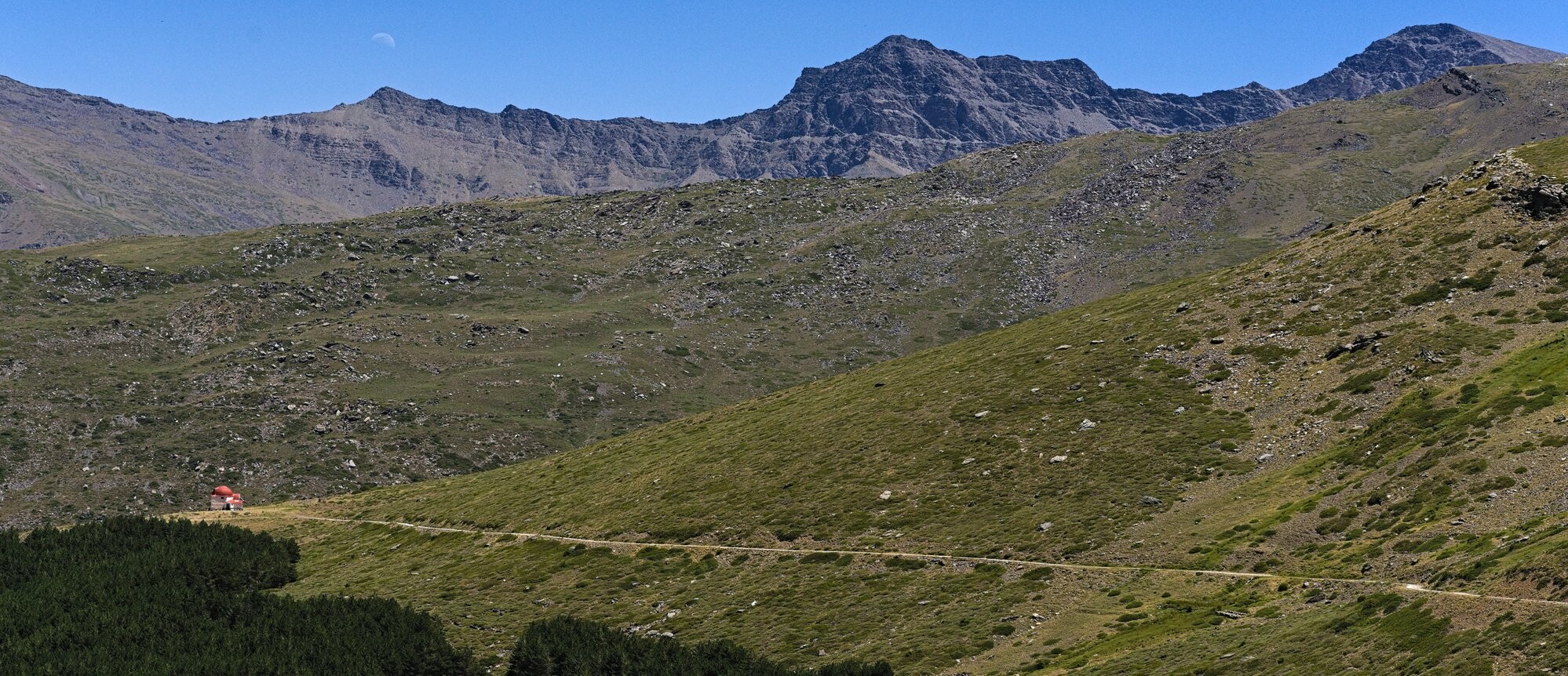 A view of the north faces of two of biggest mountains in Spain's Sierra Nevada national park. The moon rising behind one mountain. At the end of a track there is a little refugio with a red roof standing alone on the green slops with the big mountains behind.