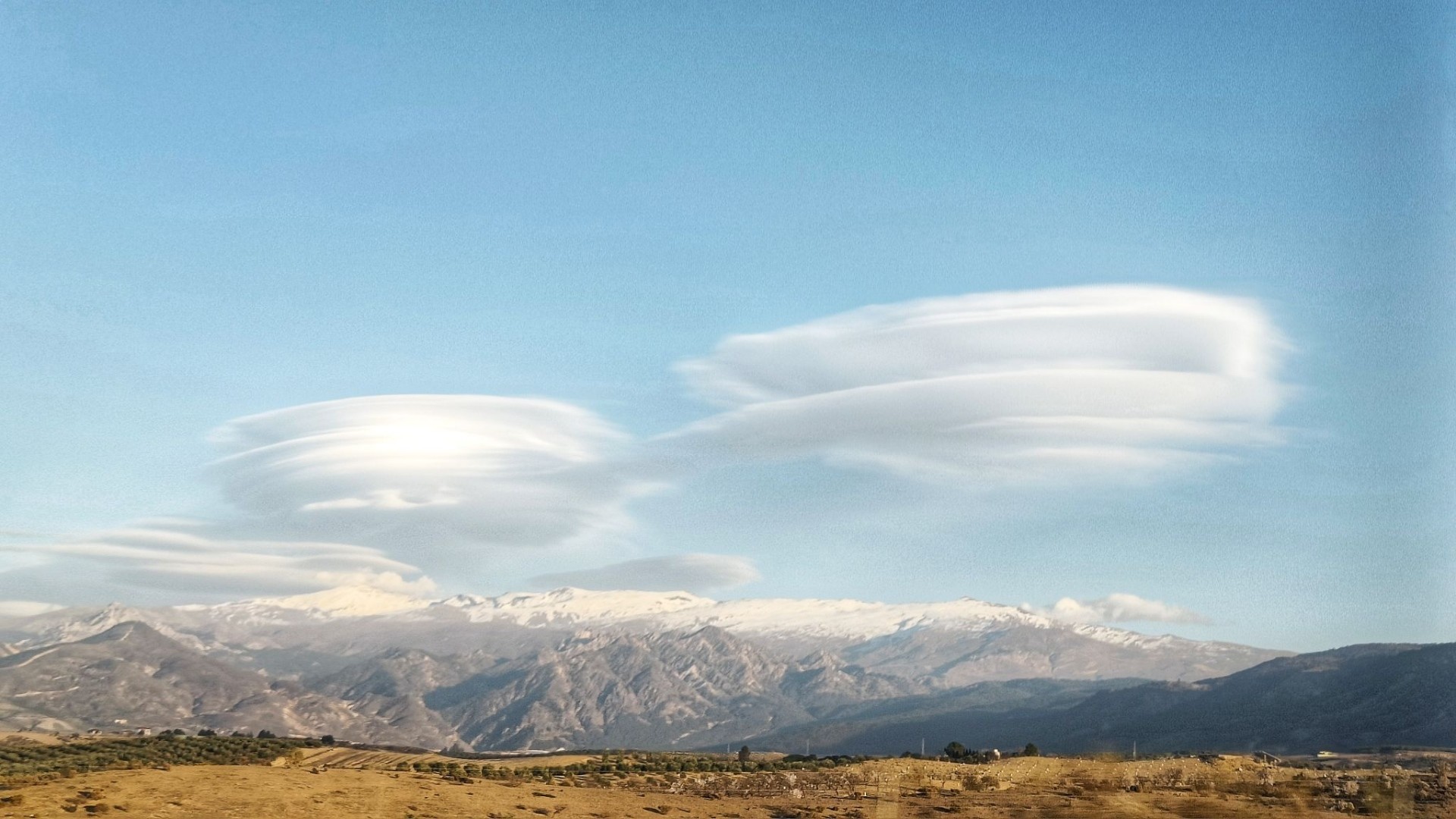 Stacked lenticular clouds in a pale blue sky above snowy mountains with lower brown mountains in front and olive trees and brown sparse ground in the forefront. Taken from a moving car.