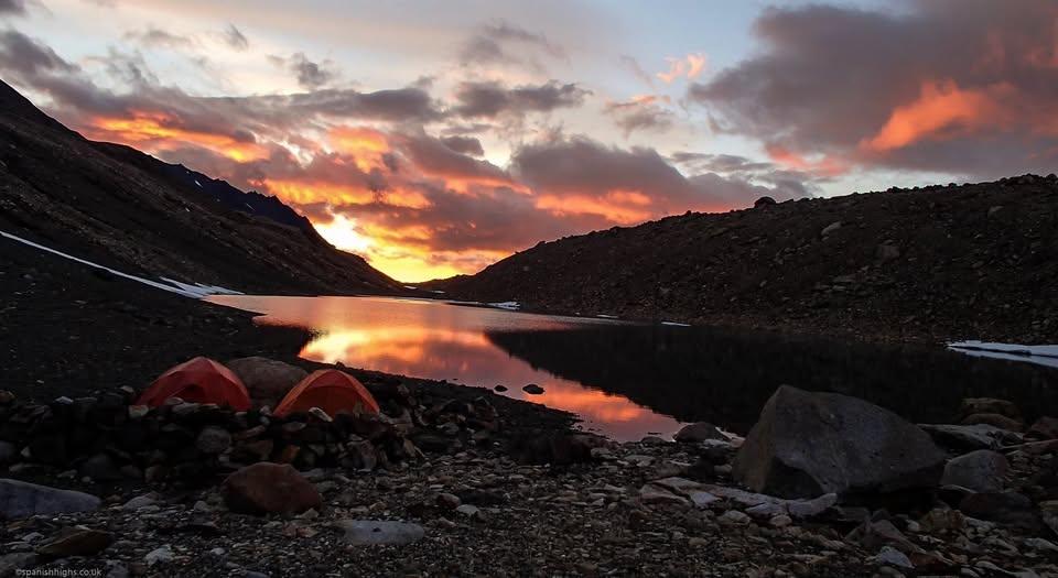 Orange sunrise reflecting on a glacial lake. The clouds and lake are orange. In the foreground are two orange tents with a rock wall around them pitched on moraine. A mountain side comes down from the left with glacial moraine on the right side of the lake. Bits of snow can be seen.