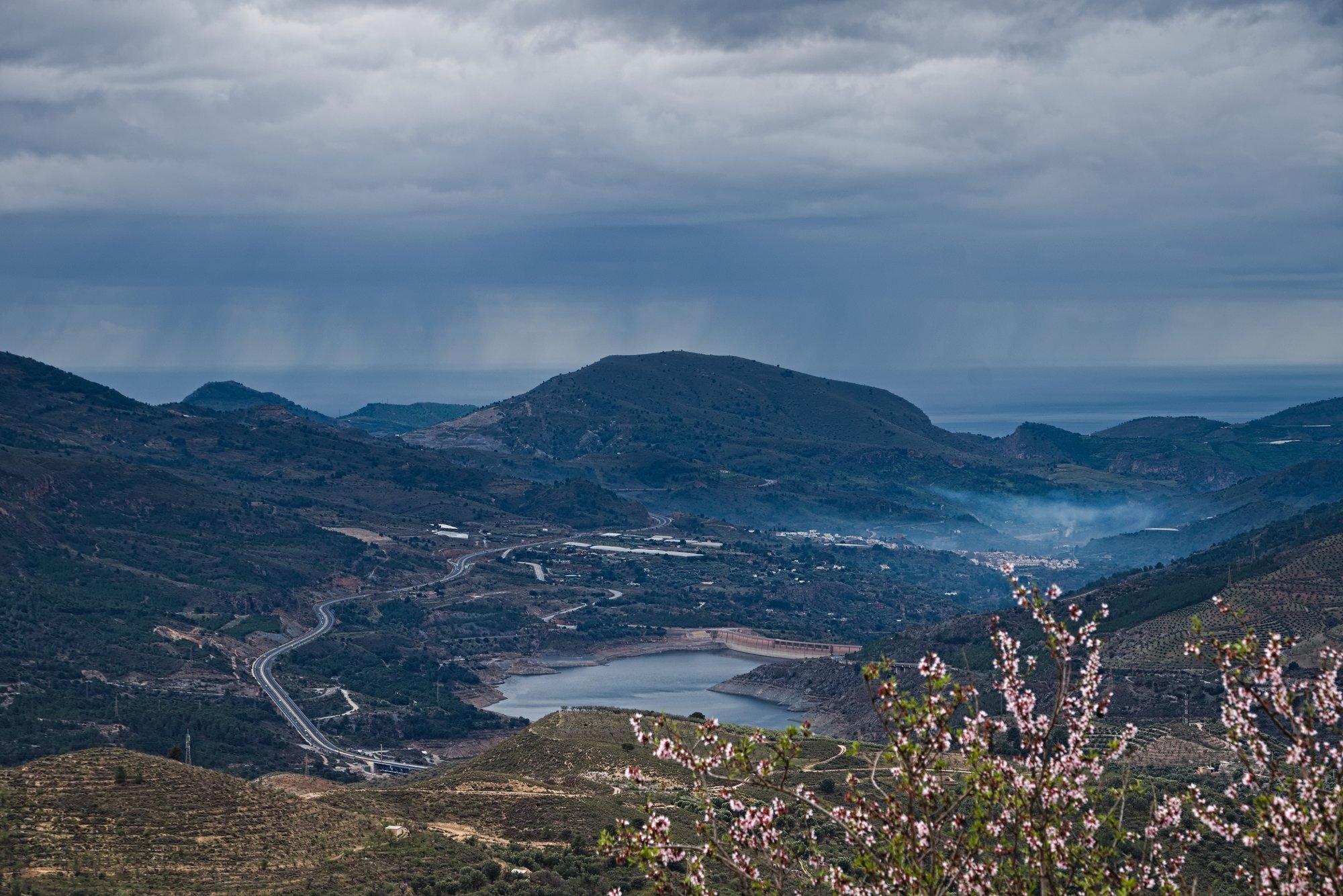 A picture showing rain falling from the sky in the distance, it looks to be falling over the Mediterranean sea or is it on the land? The sky is various shades of grey. There is an almond tree in flower in the bottom right of the picture. Various heights of hills around and a reservoir in the middle.