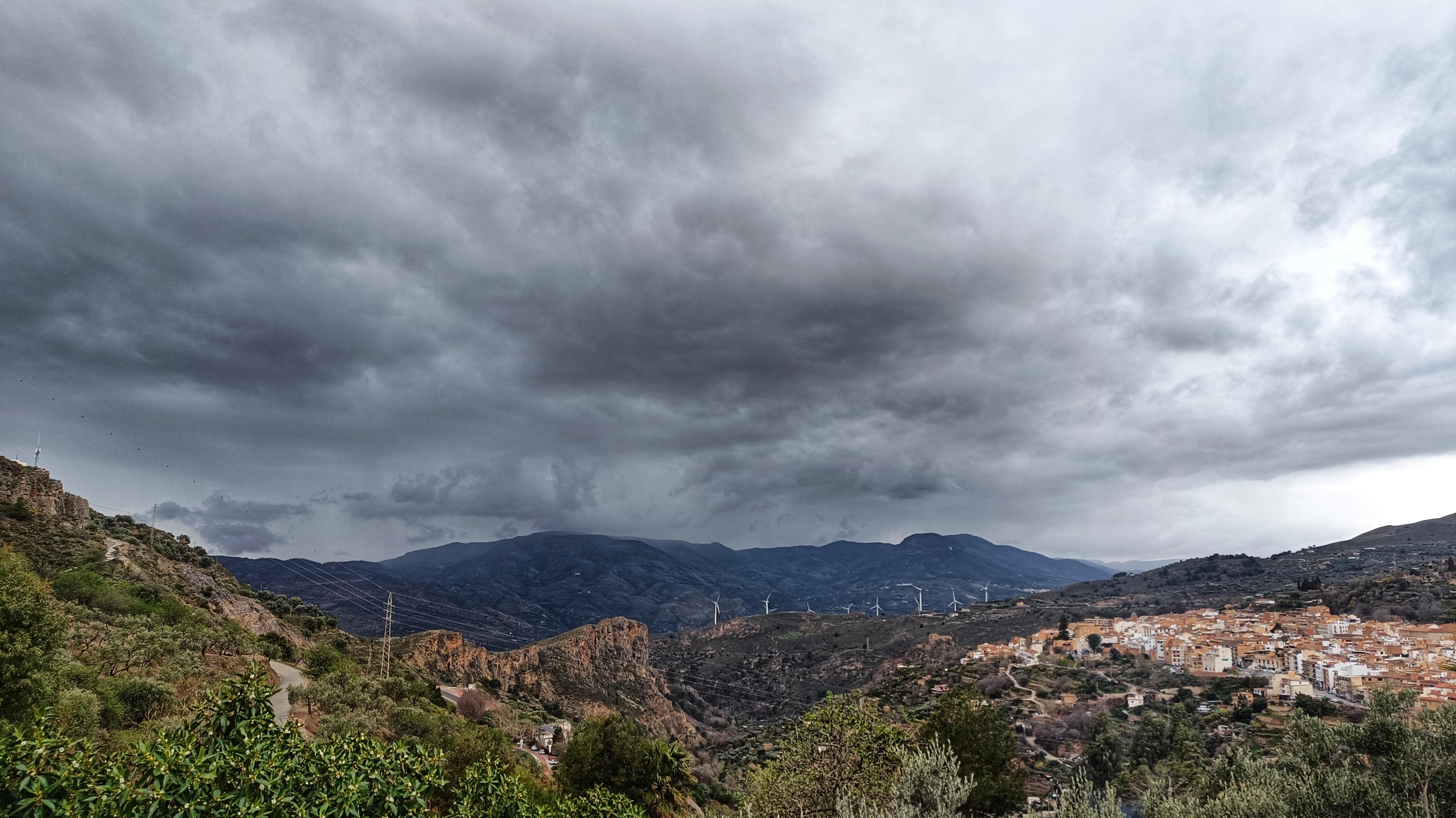 A shot looking out over the tops of trees, village on the right, towards hills in the distance. The incoming weather is coming from behind the hills. The clouds are heading towards the photographer.