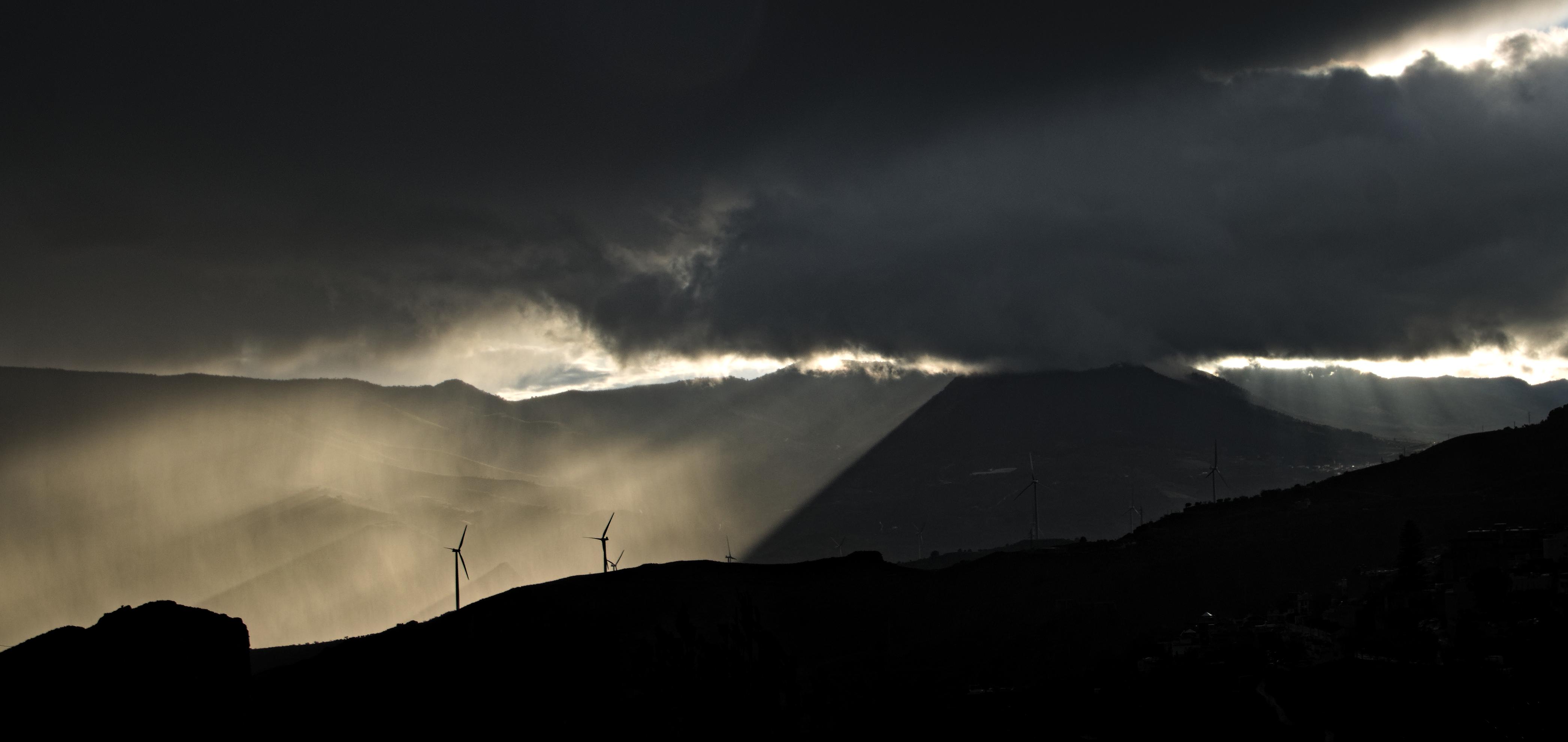 A stormy scene. Wind turbines silhouetted on a dark ridge line by a sun lit shower behind them on the left of the picture. On the right suns rays come through the clouds. Dark clouds are above. 