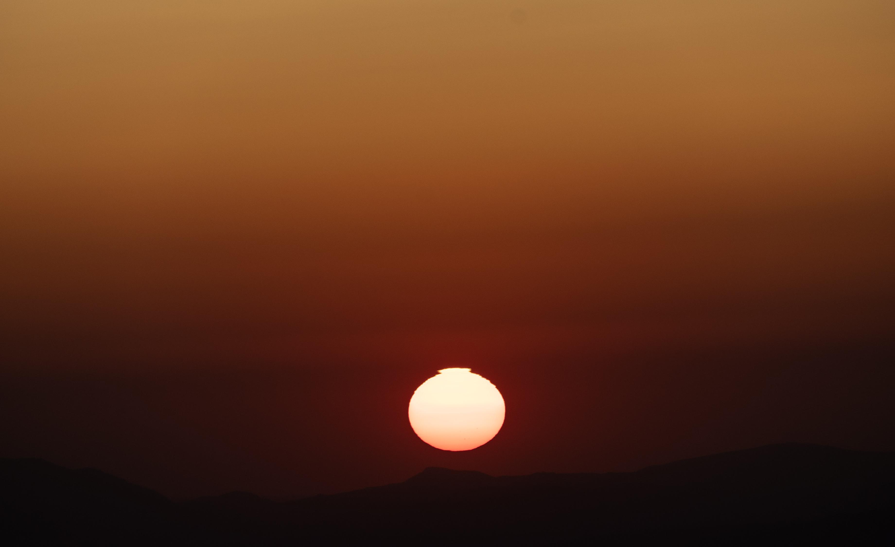 A picture of the sun setting. the sun is about to disappear below the silhouette of the mountains. The sky is various shades of orange.