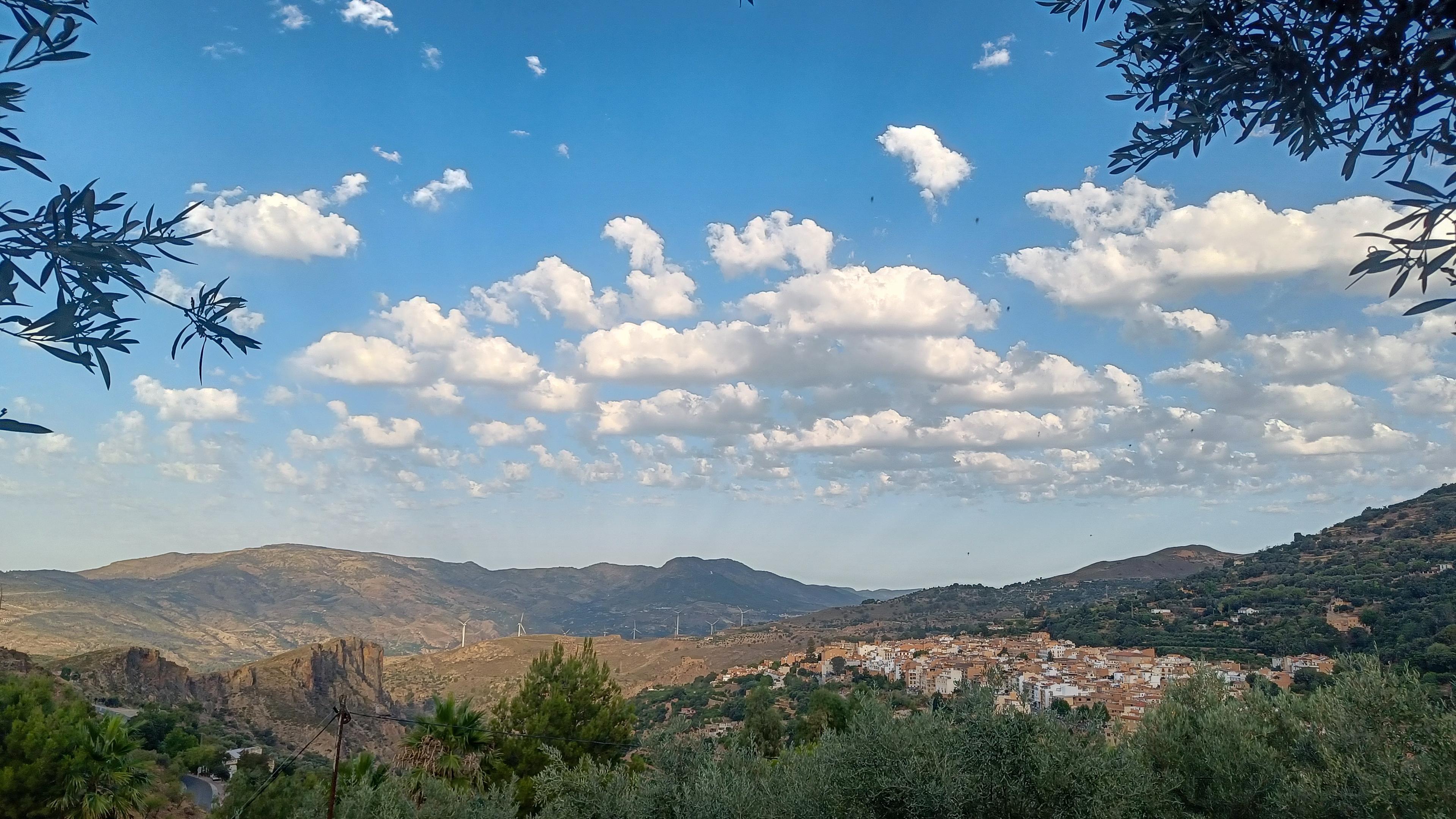 Picture showing a Spanish village. In the foreground are olive trees and behind are hillsides. The sky is various shades of blue with white cartoonish clouds.