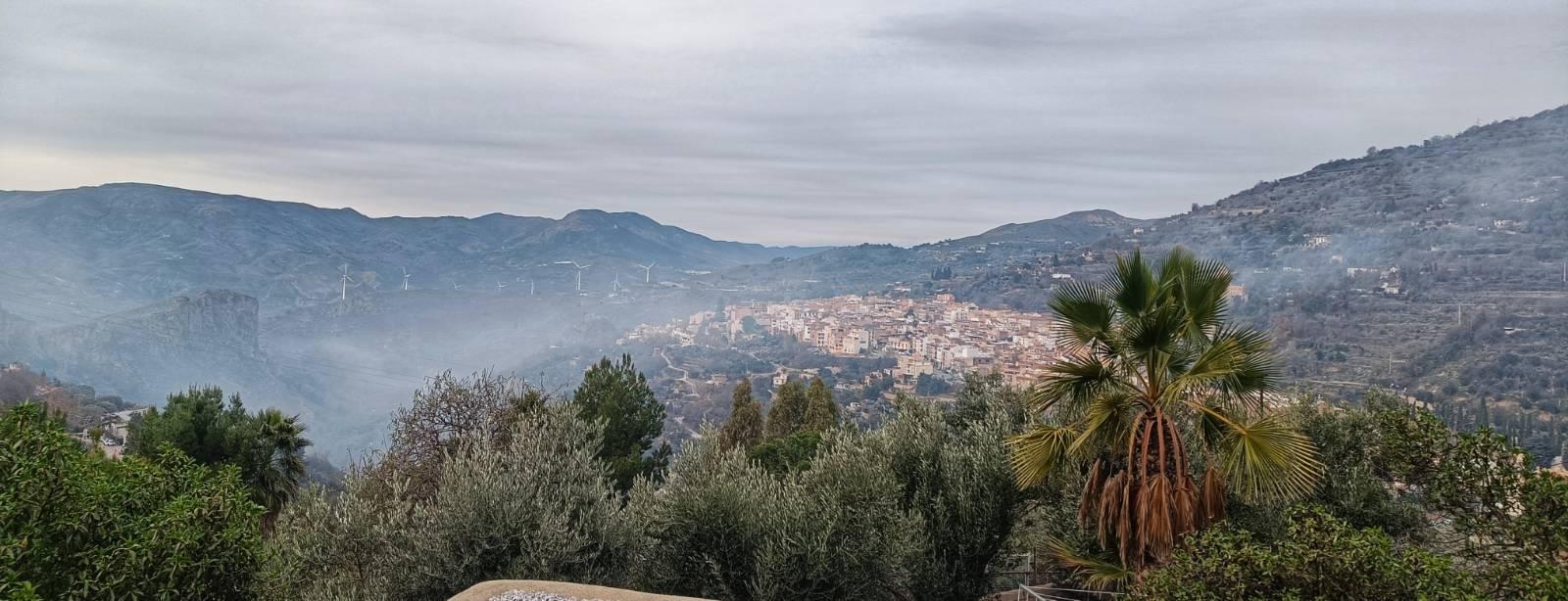 Picture showing a Spanish village, disappearing in smoke, surrounded by hillsides with green trees in the foreground. Grey cloudy sky.